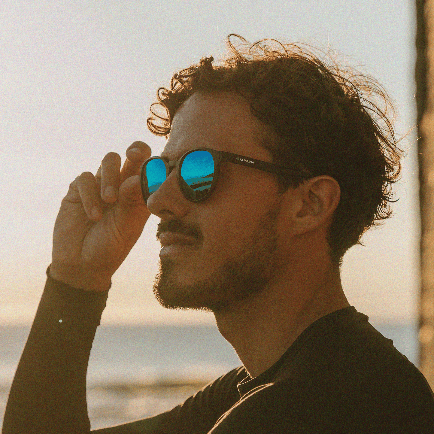 Stylish man in Kukuna® Hope Cove sunglasses with green lenses, enjoying a scenic beach view at sunset.