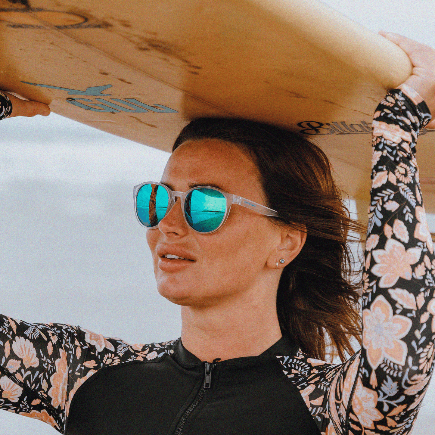 Woman in TR90 sunglasses with green mirror lens holds a surfboard, showcasing Kukuna® Hope Cove stylish performance.