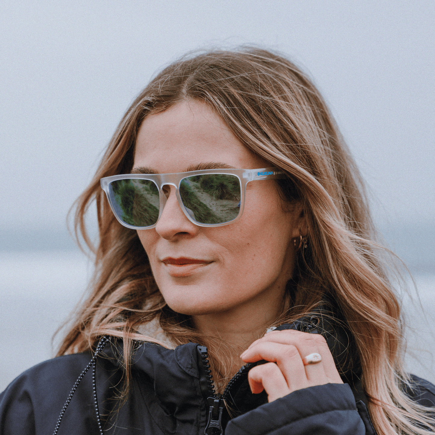 Female surfer wearing Kukuna Barricane durable sports glasses with clear frosted frame and blue mirror lenses
