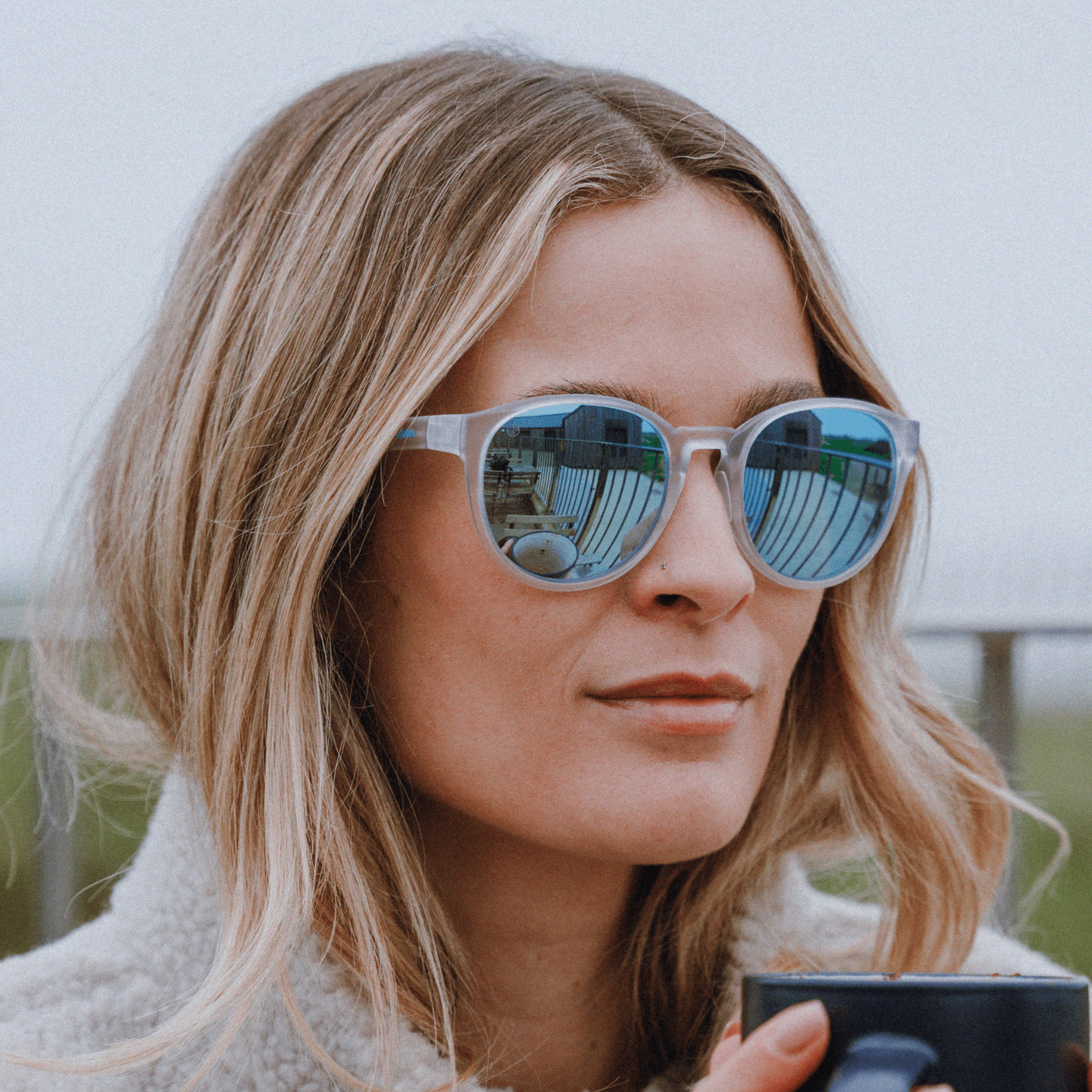 Woman wearing Kukuna® Hope Cove lifestyle sunglasses with blue mirror lenses, holding a mug in a cozy setting.