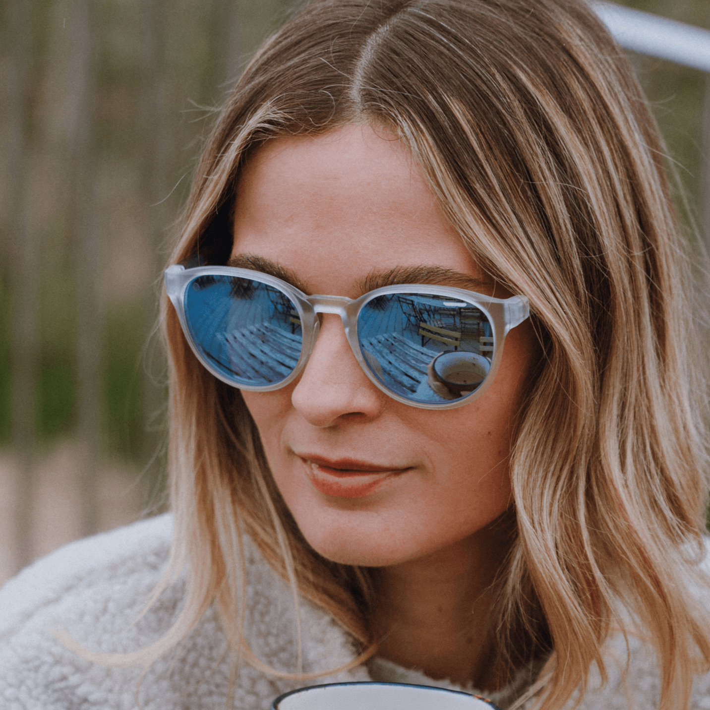 Woman wearing Kukuna® Hope Cove grey lens sunglasses with blue mirror, enjoying coffee outdoors in a cozy sweater.