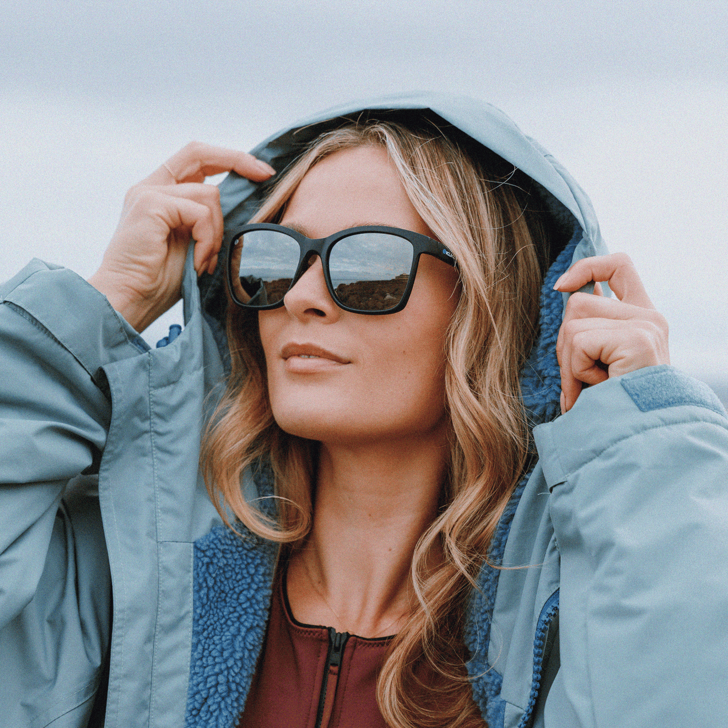 Woman wearing Kukuna Seaton lifestyle sunglasses with grey lenses and a blue hooded jacket outdoors.