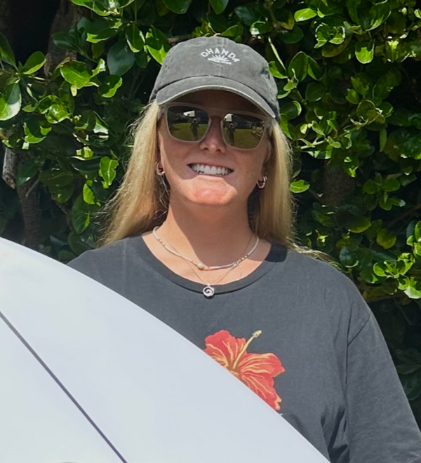 Charlotte Mulley holding a surfboard with  Kukuna branding in front of a stone wall and greenery