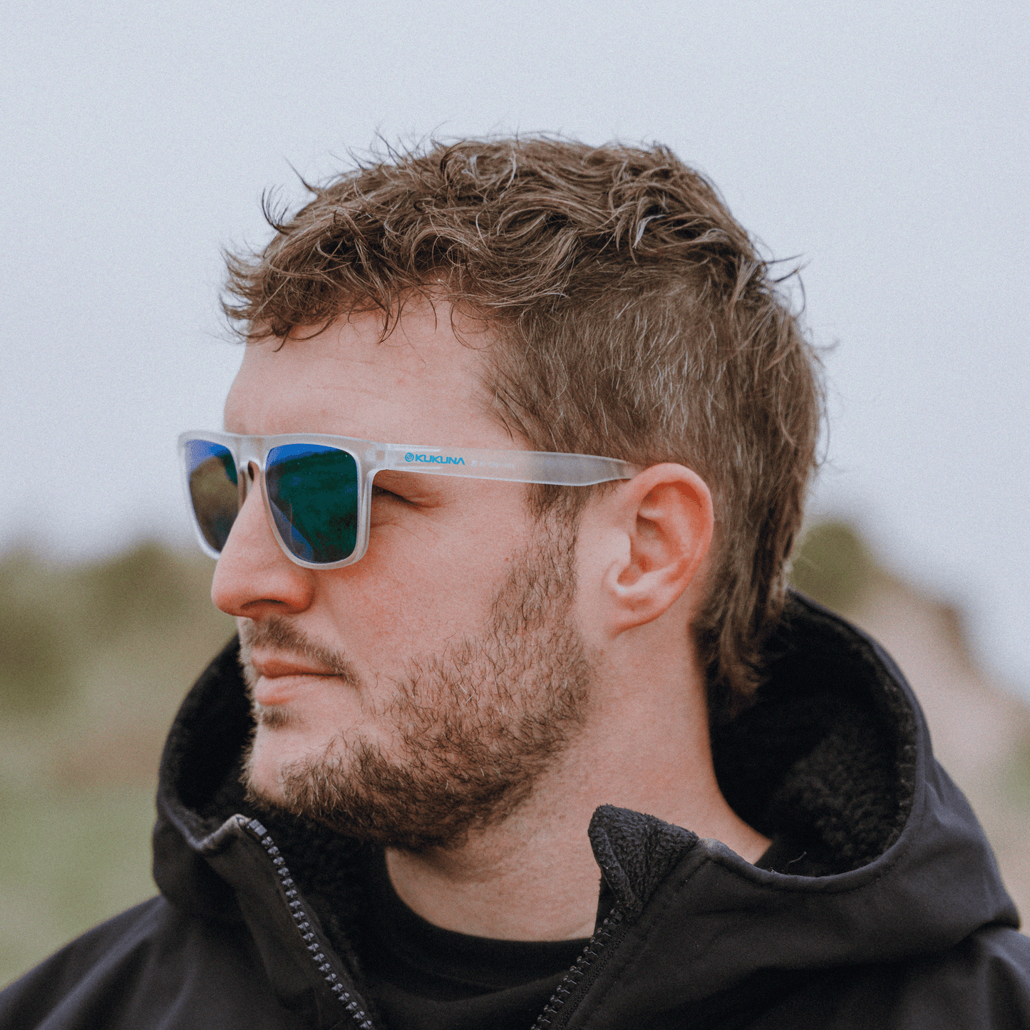 male surfer wearing  Kukuna Barricane polarised sunglasses with clear frosted frame and green mirror lenses