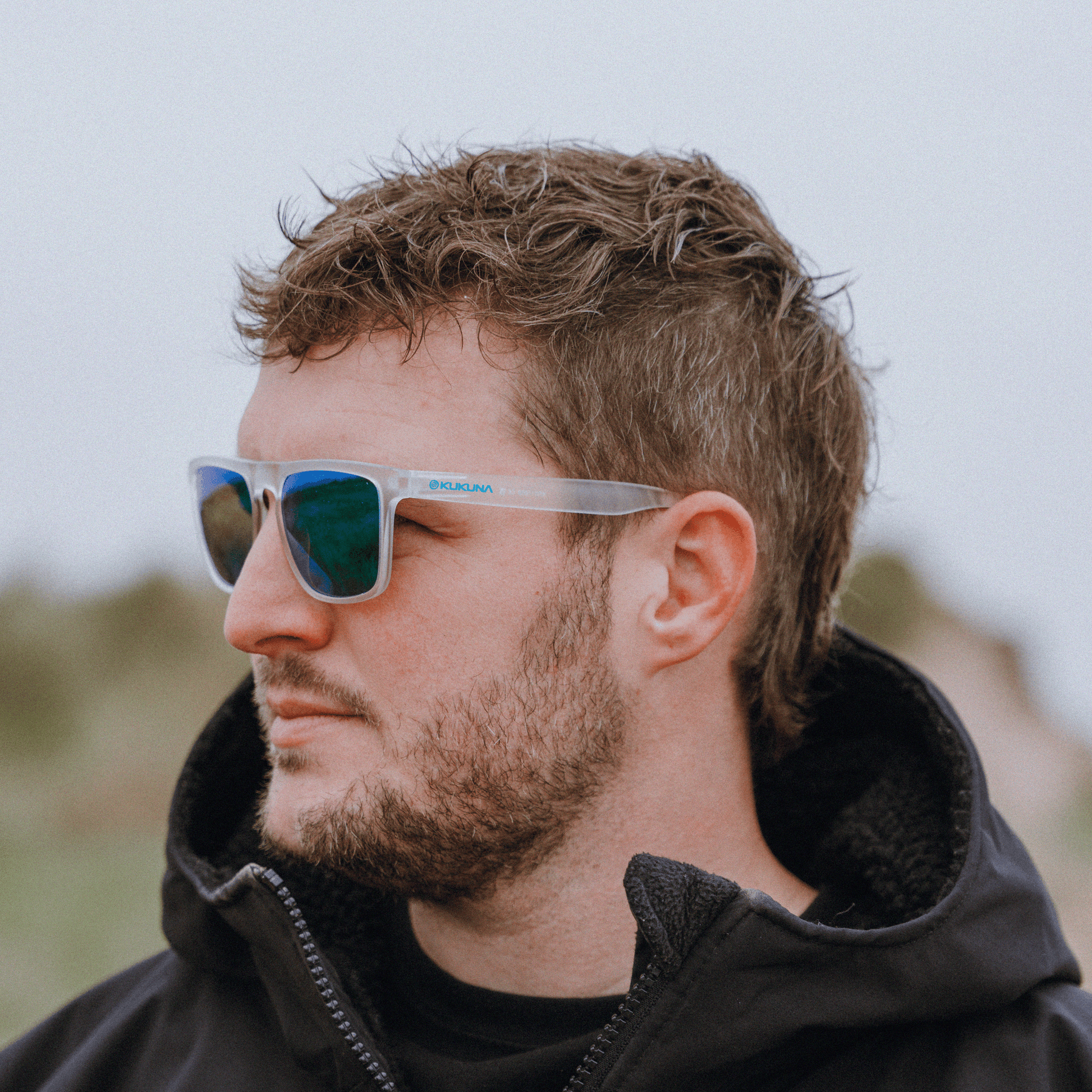 male surfer wearing  Kukuna Barricane polarised sunglasses with clear frosted frame and green mirror lenses