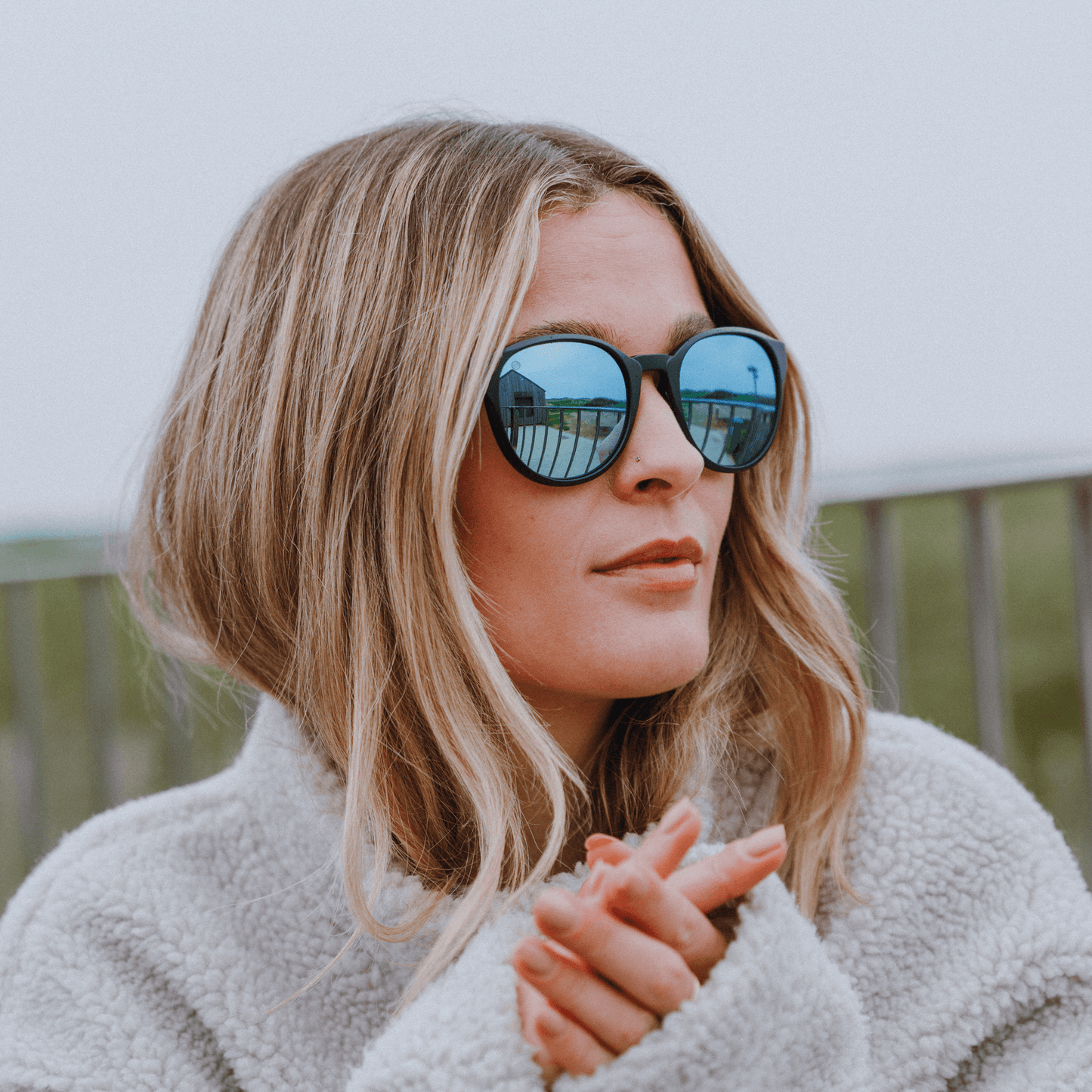Woman wearing Kukuna® Hope Cove lifestyle sunglasses with blue mirror lenses and a cozy sweater against a scenic background.