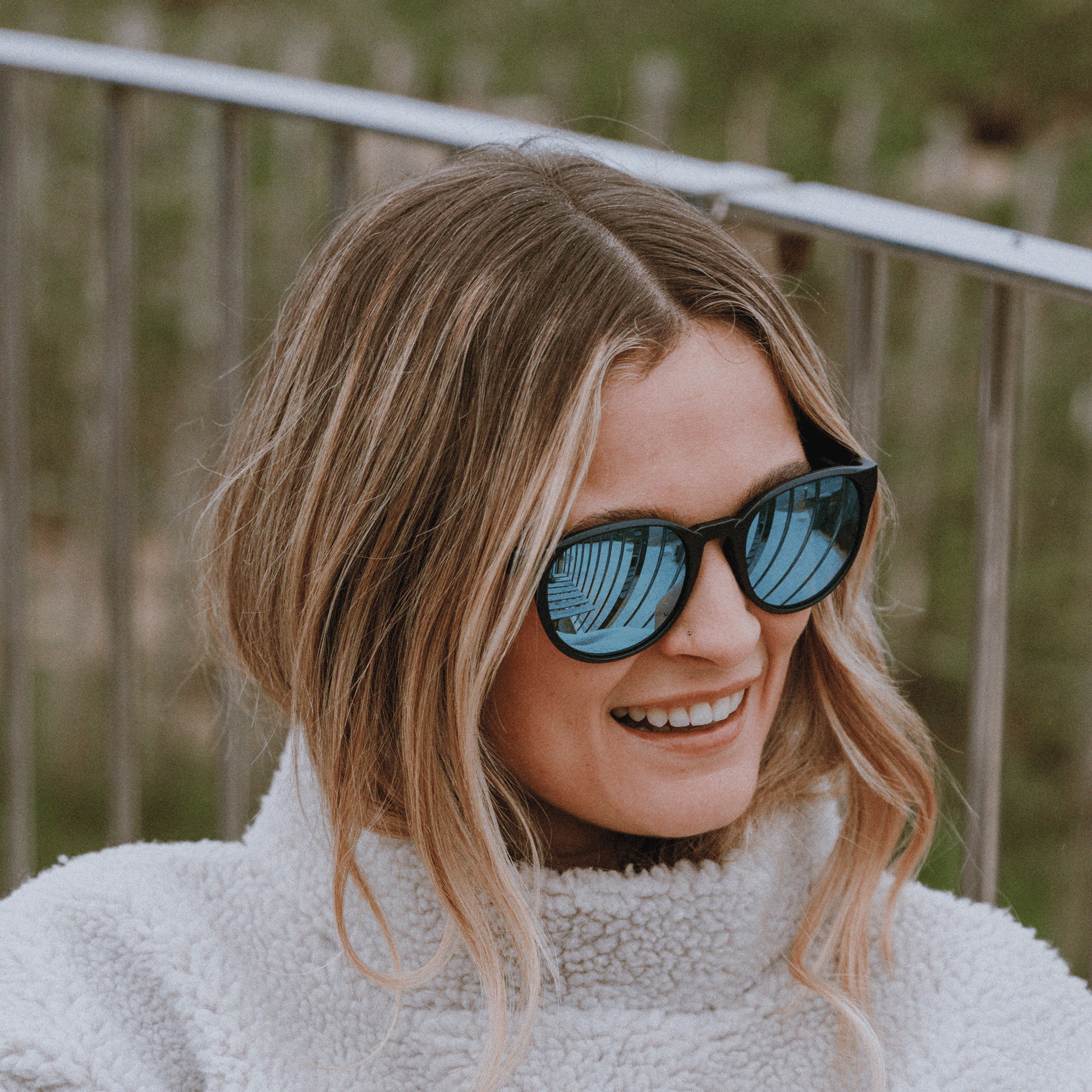 Woman wearing Kukuna® Hope Cove lifestyle sunglasses with blue mirror lenses and a cozy fleece jacket outdoors.