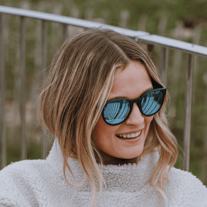 Woman wearing Kukuna® Hope Cove lifestyle sunglasses with blue mirror lenses and a cozy fleece jacket outdoors.