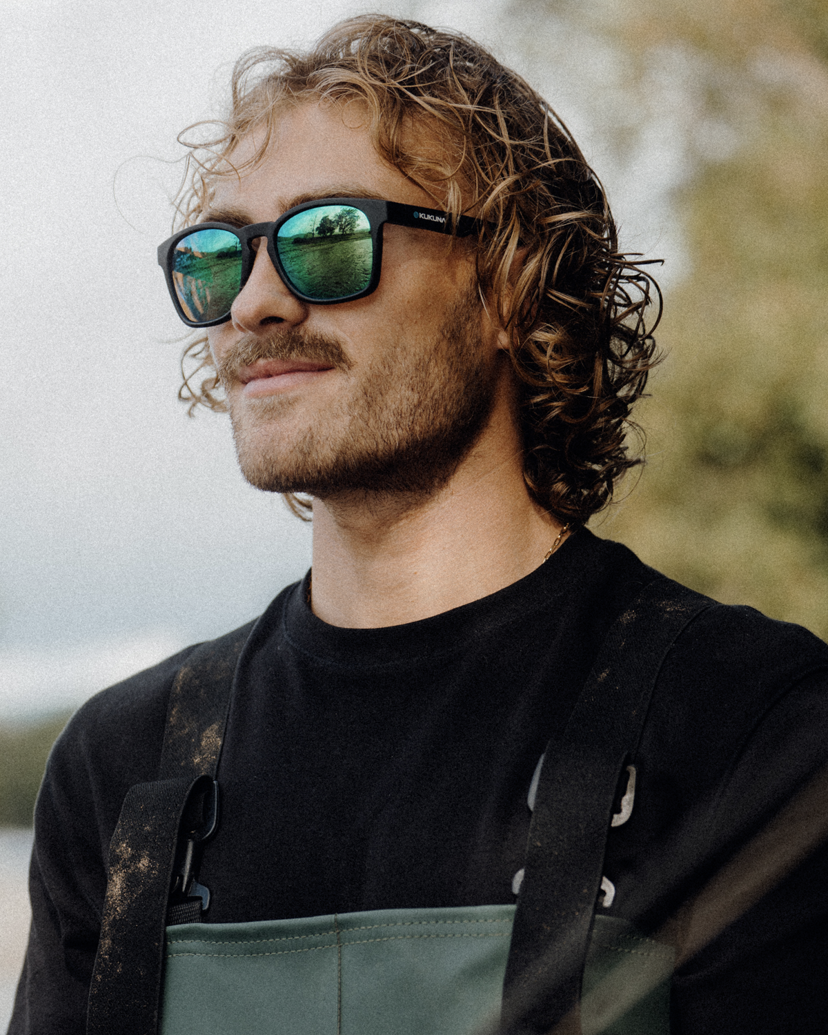 A man with curly light brown hair and a beard wearing black Kukuna sunglasses with green mirrored lenses and a black shirt, photographed outdoors against a soft blurred background.